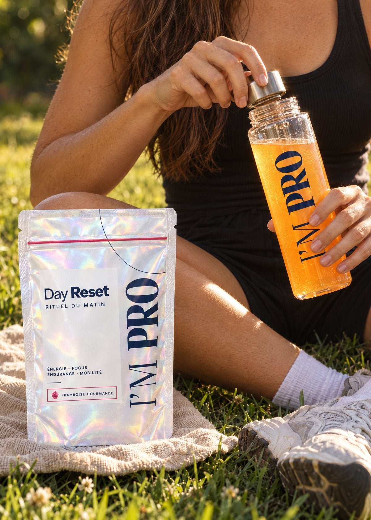Une sportive, assise dans l’herbe, ouvre sa gourde I’M PRO près de son sachet de Day Reset.