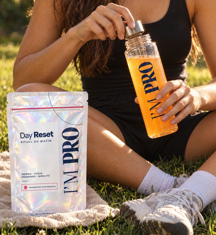 Assise dans l’herbe, une sportive ouvre sa gourde I’M PRO à côté d’un sachet de Day Reset.